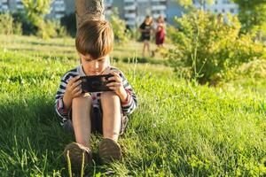 en skön barn Sammanträde på de gräs spelar på de telefon i en spel i de sommar på solnedgång. pojke har roligt i natur foto