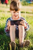 en stilig pojke Sammanträde på de gräs spelar i de telefon i de sommar spel på solnedgång. de barn har roligt i natur foto