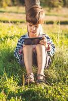 en stilig pojke Sammanträde på de gräs spelar i de telefon i de sommar spel på solnedgång. de barn har roligt i natur foto