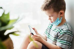 en söt barn i en medicinsk mask sitter på Hem i karantän förbi de fönster med en telefon i hans händer och utseende in i Det. förebyggande av coronavirus och covid - 19 foto