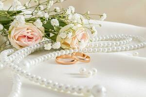 två guld bröllop ringar och underbar pärla pärlor mot de bakgrund av en bukett med ro på en beige satin bakgrund. främre se. vykort. foto