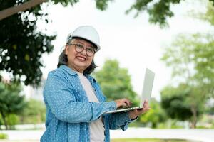 Lycklig äldre asiatisk professionell ingenjör bär en hjälm och checkar de plan på en läsplatta på de konstruktion webbplats efter de entreprenör och arkitekt Avsluta renovering de hus och byggnad foto