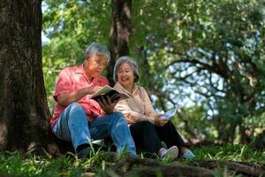 Lycklig asiatisk gammal senior par med grå hår läsning en bok utanför i parkera. begrepp av Lycklig äldre efter pensionering och Bra hälsa foto