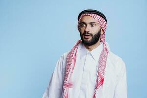 man bär traditionell muslim kläder ser bort chockade ansikte uttryck. arab person klädd i thobe och slöja stående med överraskad känsla och öppen mun medan Framställ i studio foto