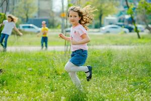 skön liten flicka med utvecklande hår promenader i gata kränkande karantän av coronavirus. flicka i rosa t-shirt kör tvärs över grön fält. flicka spelar aktiva spel i natur i sommar eller vår. foto