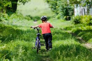 en pojke bär hans cykel. barn i en skyddande hjälm går längs med hans cykel. en liten pojke kommer tillbaka från en promenad genom de by. foto