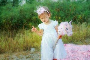 liten skön flicka i en vit klänning Hoppar för unicorns på en pinne. de flicka i de krona lutande hans huvud och pekande hans finger. en barnets födelsedag är på de gata. foto