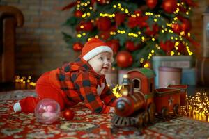 liten flicka Nästa till jul tåg. Lycklig barn i santa hatt nära gåvor på bakgrund av ny år träd i festlig rum. 6 månad gammal bebis är leende. skön liten flicka väntar för jul presenterar. foto