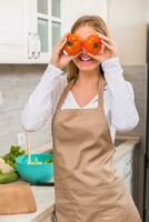 skön kvinna beläggning ögon med tomat medan framställning måltid i henne kök foto