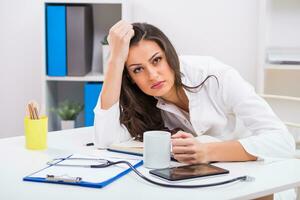 trött kvinna läkare är dricka kaffe medan arbetssätt på henne kontor foto