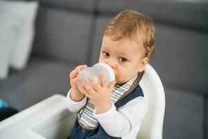 söt bebis pojke dricka vatten medan Sammanträde i en hög stol foto