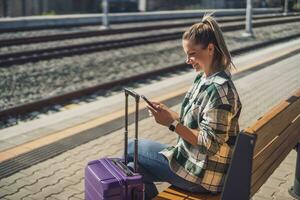 Lycklig kvinna använder sig av mobil telefon medan Sammanträde på en bänk på de tåg station foto