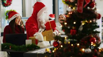 multietnisk arbetare tar emot gåvor från kollega låtsas till vara santa i xmas dekorerad kontor. anställd bär kostym förvånande företag personal med presenterar under vinter- Semester säsong foto