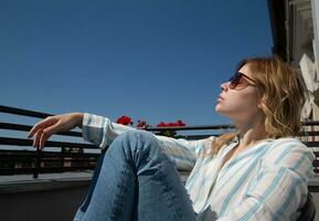 skön flicka i solglasögon sitter i en stol på de balkong i de morgon- ljus mot de himmel. hög kvalitet Foto