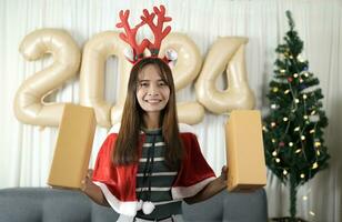 asiatisk kvinna innehav en gåva låda ha roligt fira jul 2024. dekorera och dekorera de rum med en jul träd och 2024 ballonger. foto