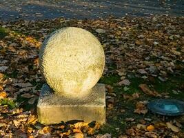 en skulptur i de form av en boll på en fyrkant piedestal i en stad parkera. de spela av ljus på de yta av de boll. foto