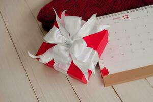 röd gåva låda och kalender på isolerat på en röd stickat scarf med vit trä- bakgrund med. julen dag begrepp. planera schemaläggning dagordning, händelse, arrangör x'mas dag. platt lägga, topp se. foto