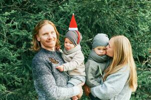 utomhus- porträtt av skön Lycklig ung familj av 4 Framställ i tall skog, bär värma tröjor, par med litet barn pojke och bebis flicka har Bra tid på natur, kall väder, jul tema foto