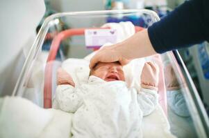 nyfödd gråt bebis i sjukhus spjälsäng, far påfrestande till lugna ner spädbarn förbi sätta hans hand åh henne huvud foto