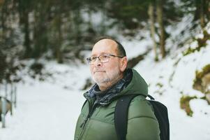 utomhus- porträtt av mitten ålder 55 - 60 år gammal man vandring i vinter- skog, bär värma jacka och svart ryggsäck foto