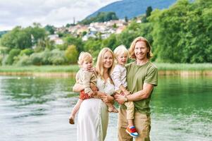 utomhus- porträtt av skön familj, ung par med preschooler pojke och litet barn flicka Framställ Nästa till sjö eller flod foto