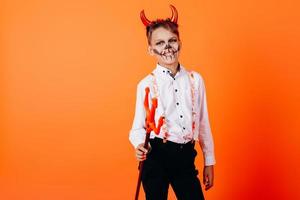 djävulen pojke står halv-turn mot en orange bakgrund i maskerad makeup. halloween koncept foto