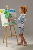 charmig skola flicka är målning med en vattenfärg borsta på ett staffli, stående på en grå bakgrund. foto