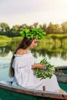 de nymf med lång mörk hår i en vit årgång klänning Sammanträde i en båt i de mitten av de flod. foto