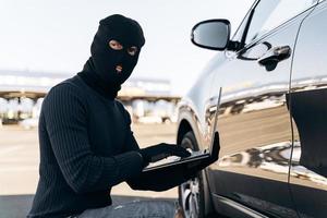 midja upp porträtt av biltjuven med larmsystem för laptophackning medan du sitter nära bilen på dagtid. lager foto