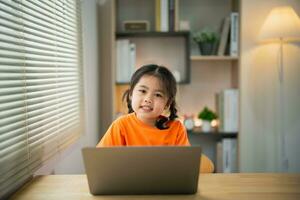 asiatisk bebis flicka bär t-shirt leende koncentrera till använda sig av bärbar dator och studie uppkopplad på trä tabell skrivbord i levande rum på Hem på natt. utbildning inlärning uppkopplad från Hem begrepp. foto