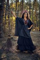 häxa i svart, lång klänning, med röd krona i henne lång hår. Framställ med kvast i tall skog. trollformler, magi och trolldom. full längd porträtt. foto