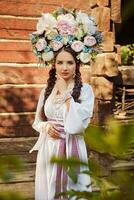 brunett flicka i en vit ukrainska äkta nationell kostym och en krans av blommor är Framställ mot en trä- hus. närbild. foto