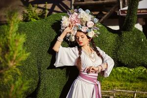 brunett flicka i en vit ukrainska äkta nationell kostym och en krans av blommor är Framställ mot en grön oxe ristade från en buske. foto