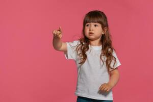 närbild porträtt av en liten brunett flicka klädd i en vit t-shirt Framställ mot en rosa studio bakgrund. uppriktig känslor begrepp. foto