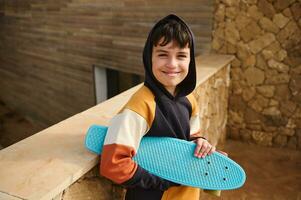 glad tonåring pojke, hipster, skateboarder i trendig luvtröja, innehav hans blå skateboard, leende ser på kamera foto