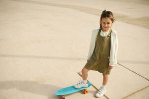 leende liten barn flicka stående på en skateboard på urban skatepark och ser upp på de kamera foto