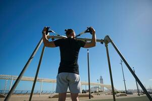 ung attraktiv atletisk man håller på med övre kropp övning Träning vapen, använder sig av suspension remmar utomhus ensam. bak- se foto