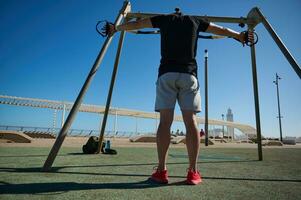 bak- se av ett attraktiv man under kroppsvikt träna med suspension remmar i de utomhus- idrottsplats. foto