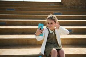 Lycklig unge flicka talande via video länk, framställning selfie, Sammanträde på steg utomhus på värma solig dag. uppkopplad kommunikation foto