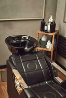 svart läder sittplats med tvätta handfat i en frisör interiör med handdukar och schampo på de sida. foto
