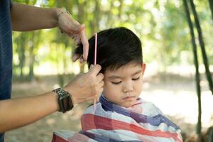 barberare skärande hår av ett asiatisk pojke i ett öppen Plats fylld med träd. foto
