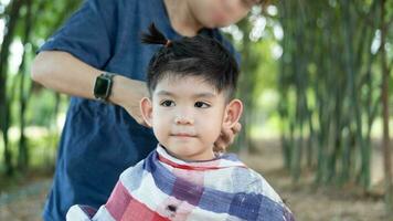 barberare skärande hår av ett asiatisk pojke i ett öppen Plats fylld med träd. foto
