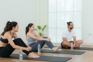 en grupp av kvinna och manlig idrottare Sammanträde resten på yoga mattor och dricka vatten. de är ser på varje Övrig och leende foto