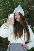 leende brunett tonåring flicka använder sig av mobiltelefon framställning video selfie till fångande stunder från tall skog foto