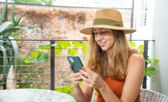 Lycklig attraktiv kvinna med hatt använder sig av smartphone leende på en terrass utanför. foto