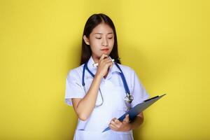 skön ung kvinna gör en omtänksam uttryck medan bär en läkares enhetlig stående i främre av en gul bakgrund. foto