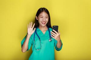 porträtt av en skön ung kvinna i en gul bakgrund, asiatisk kvinna poser med en cell telefon medan bär en läkares enhetlig och en stetoskop. foto