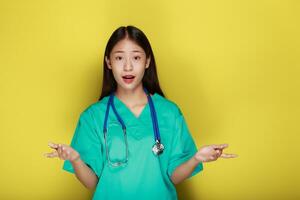 ett asiatisk kvinna bär en läkares enhetlig poser med händer medan förklara, stående i främre av en gul bakgrund. foto