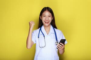 porträtt av en skön ung kvinna i en gul bakgrund, asiatisk kvinna poser med en cell telefon medan bär en läkares enhetlig och en stetoskop. foto