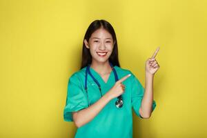porträtt av en skön ung kvinna i en gul bakgrund, vänlig skön kvinna bär en läkares enhetlig och pekande medan bär en stetoskop. foto